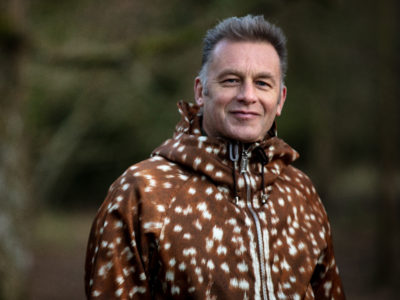 Chris Packham looking at the camera against a green blur background wearing a brown coat with white spots
